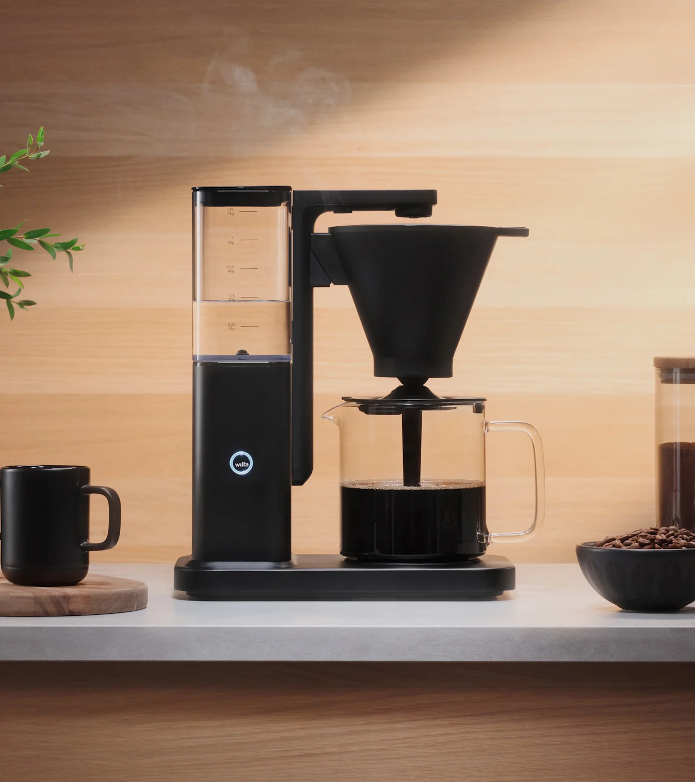 En sort kaffemaskine brygger kaffe i en glaskaraffel på en lys disk, med damp der stiger op. En sort kop og kaffebønner er i nærheden.