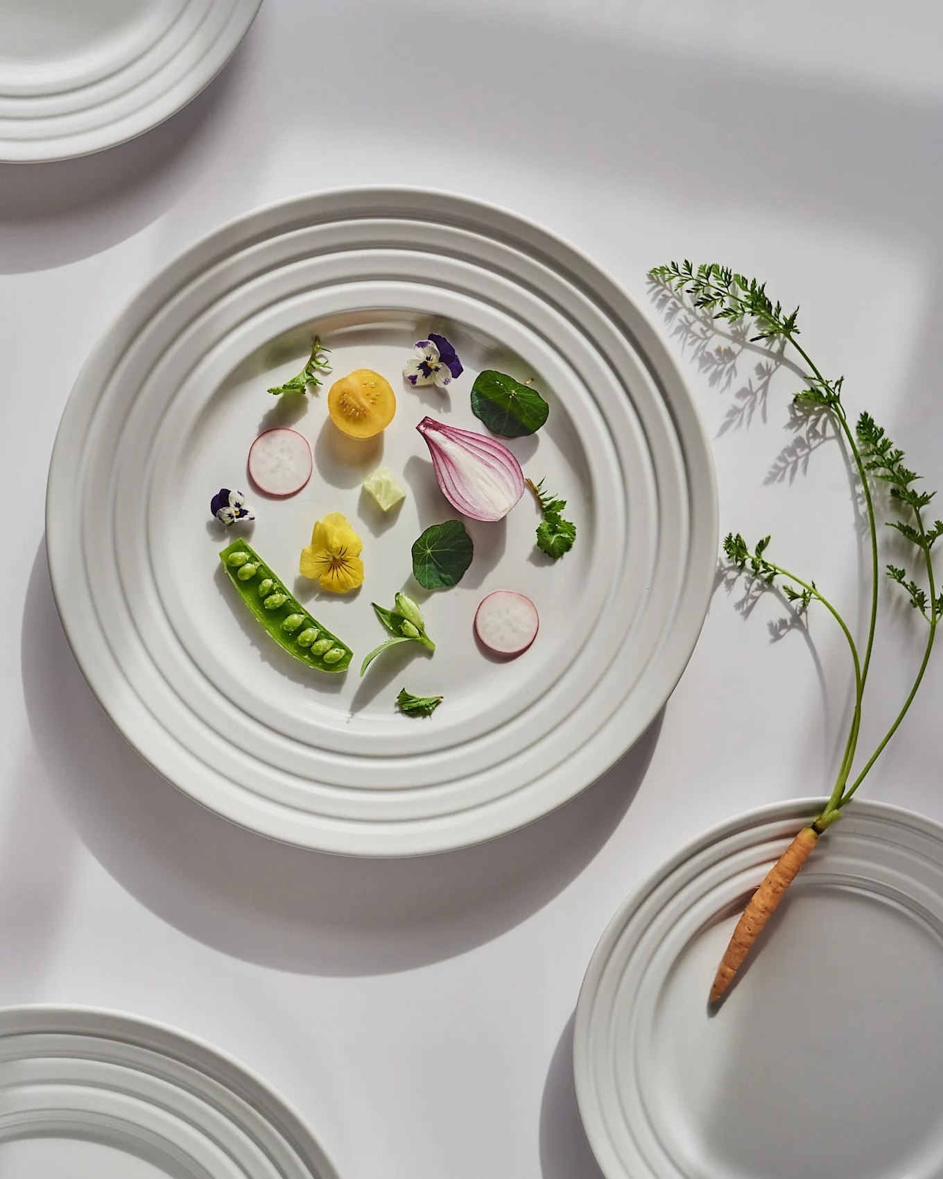 En hvid tallerken viser friske grøntsager og spiselige blomster, med en hel gulerod og dens top på en anden tallerken.