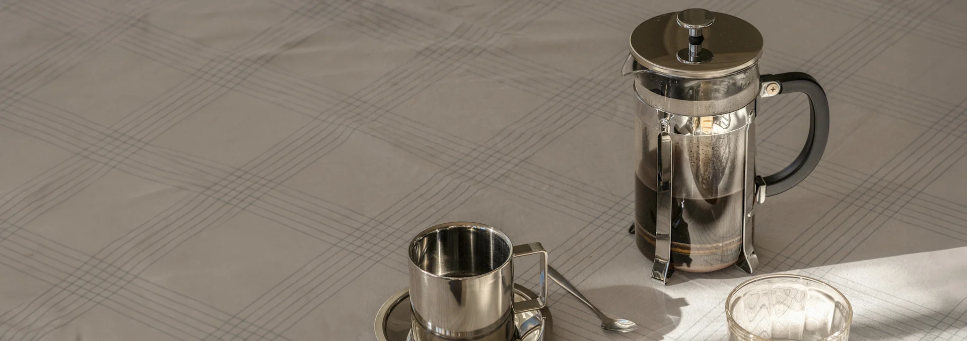 Fransk stempelkande med kaffe, stålkop, underkop og ske på et lyst, ternet lagen i sollys.