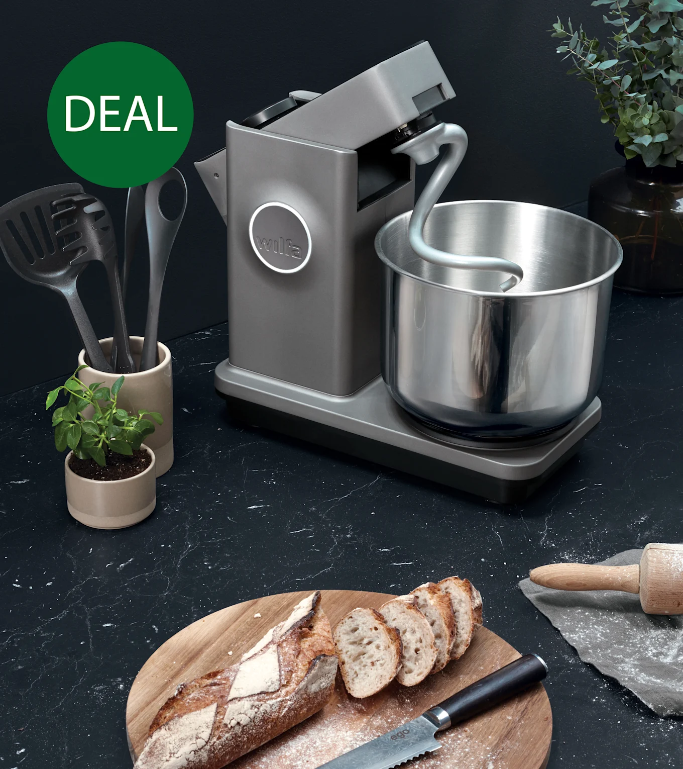 Grå køkkenmaskine med dejkrog og skål i rustfrit stål på mørk bordplade med skåret brød, markeret med grøn 'TILBUD'-cirkel.